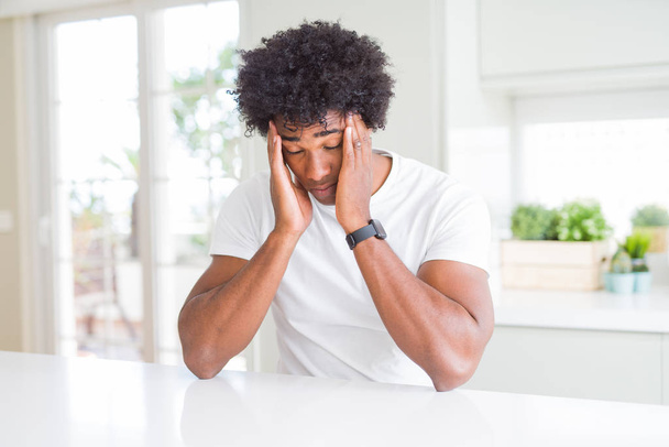 Jeune homme afro-américain portant un t-shirt blanc décontracté assis à la maison avec la main sur les maux de tête parce que le stress. Migraine souffrante
. - Photo, image