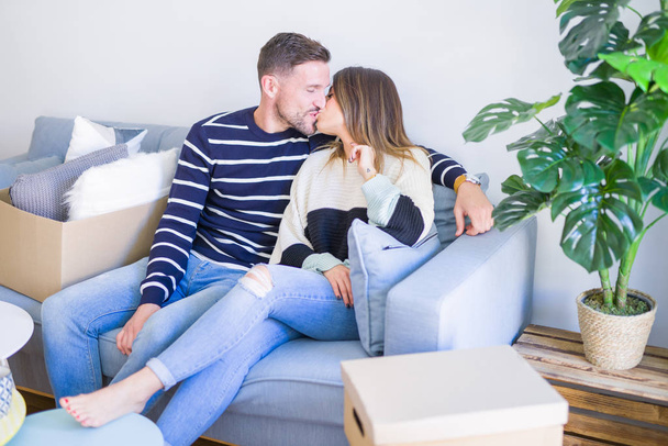 Young beautiful couple sitting on the sofa drinking cup of coffee and kissing at new home around cardboard boxes - Фото, изображение