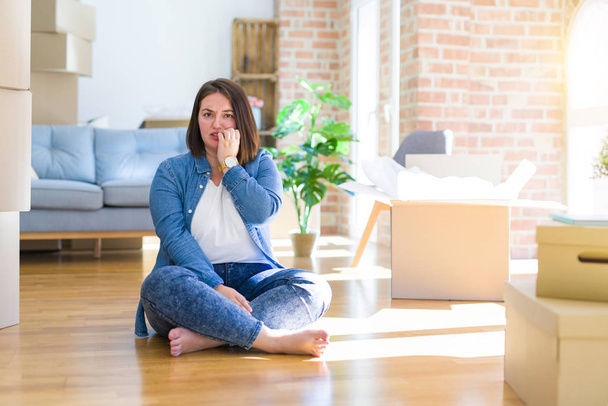 Young plus size woman sitting on the floor around cardboard boxes moving to a new home looking stressed and nervous with hands on mouth biting nails. Anxiety problem. - Photo, Image