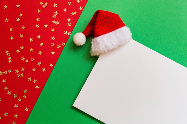Blank of paper with Santa hat on red and green background with confetti. Christmas and new year concept. Greeting card, xmas celebration 2020. Flat lay, template, top view, copy space, mockup - Foto, Imagem