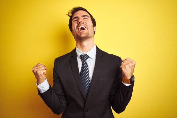 Young handsome businessman wearing suit and tie standing over isolated yellow background celebrating surprised and amazed for success with arms raised and eyes closed. Winner concept. - Foto, immagini