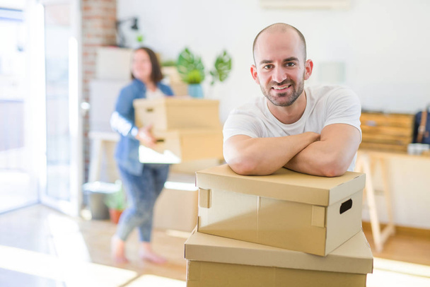 Νεαρό ζευγάρι μετακομίζει σε νέο διαμέρισμα, όμορφος άντρας που γέρνει σε χαρτοκιβώτια και χαμογελαστός χαρούμενος - Φωτογραφία, εικόνα