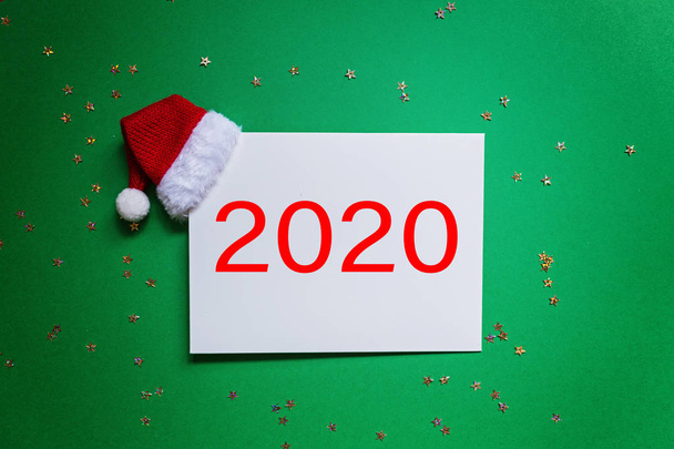 Blank of paper with red Santa hat on green background with confetti. new year concept. Greeting card, xmas celebration 2020. Flat lay, template, top view, copy space, mockup - Foto, Imagen