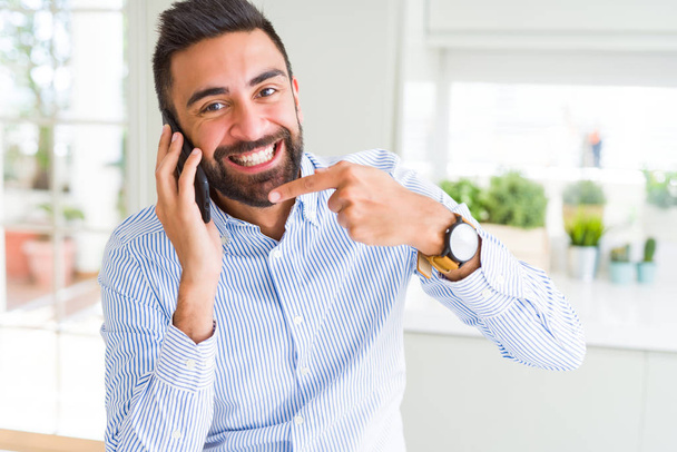 Komea latinalaisamerikkalainen liikemies, jolla on keskustelu älypuhelimella, erittäin onnellinen osoittaen kädellä ja sormella
 - Valokuva, kuva