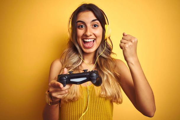 Mladá krásná žena hrající videohru pomocí Gamepad přes žluté izolované pozadí křičící a oslavující vítězství a úspěch velmi vzrušené, povzbuzující emoce - Fotografie, Obrázek