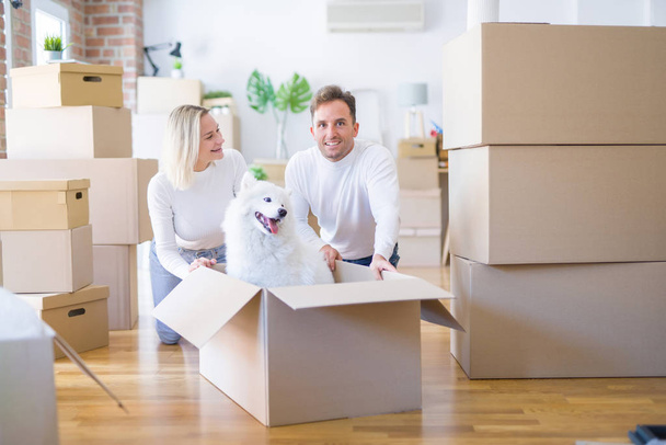 Young beautiful couple with dog sitting on the floor at new home around cardboard boxes - Фото, изображение