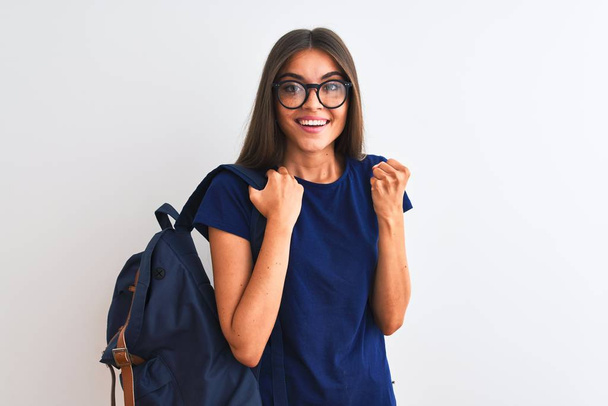 Young beautiful student woman wearing backpack and glasses over isolated white background screaming proud and celebrating victory and success very excited, cheering emotion - 写真・画像