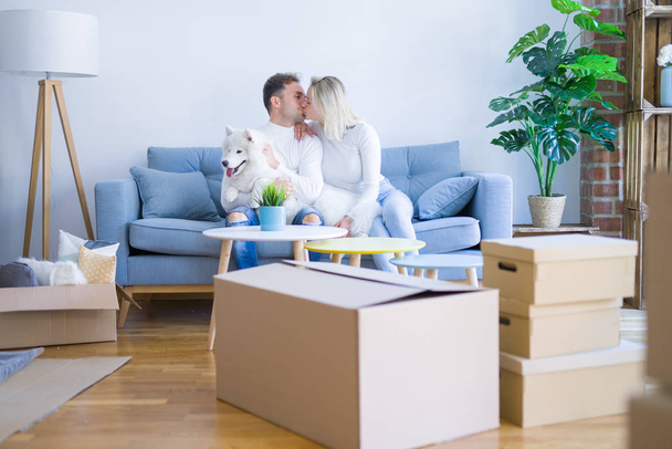 junges schönes Paar mit Hund küsst auf dem Sofa im neuen Zuhause um Kartons herum - Foto, Bild