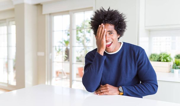 Young african american man wearing casual sweater sitting at home covering one eye with hand, confident smile on face and surprise emotion. - Fotografie, Obrázek