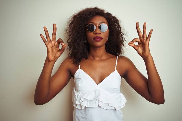 Mladá Afroameričanka v tričku a slunečních brýlích na izolovaném bílém pozadí odpočívá a usmívá se se zavřenýma očima a prsty provádí meditační gesto. Koncept jógy. - Fotografie, Obrázek