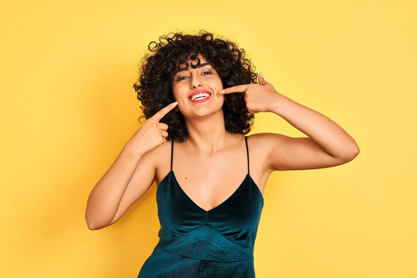 Young arab woman with curly hair wearing elegant dress over isolated yellow background smiling cheerful showing and pointing with fingers teeth and mouth. Dental health concept. - Fotoğraf, Görsel