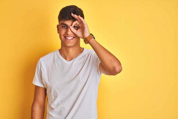 Young indian man wearing white t-shirt standing over isolated yellow background doing ok gesture with hand smiling, eye looking through fingers with happy face. - Valokuva, kuva