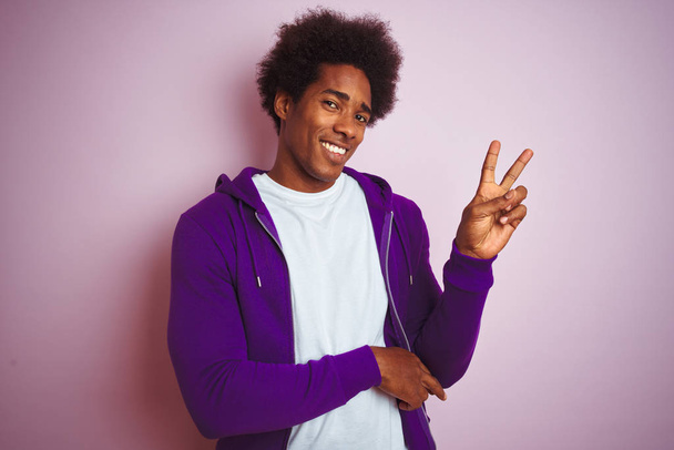 Jovem afro-americano vestindo camisola roxa em pé sobre fundo rosa isolado sorrindo com o rosto feliz piscando para a câmera fazendo sinal de vitória com os dedos. Número dois.
. - Foto, Imagem