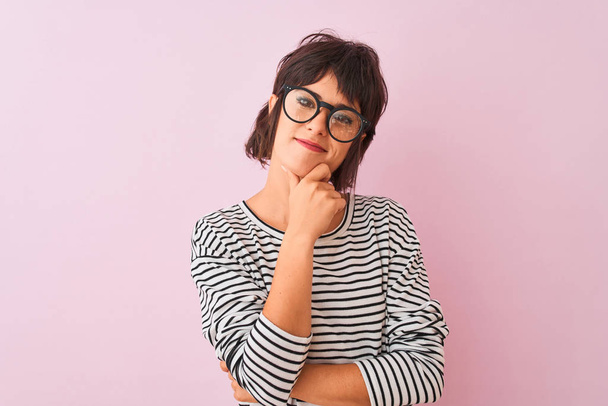 Young beautiful woman wearing striped t-shirt and glasses over isolated pink background looking confident at the camera smiling with crossed arms and hand raised on chin. Thinking positive. - Photo, Image