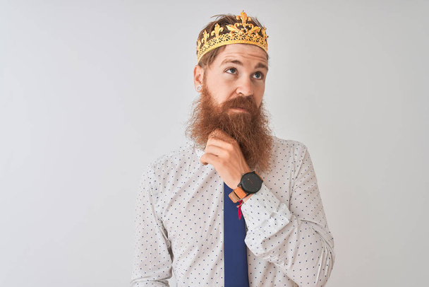 Young redhead irish businessman wearing crown king over isolated white background with hand on chin thinking about question, pensive expression. Smiling with thoughtful face. Doubt concept. - Фото, изображение