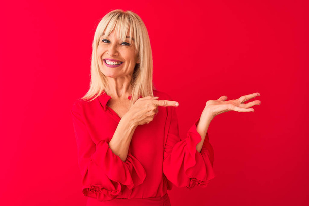 Middle age woman wearing elegant shirt standing over isolated red background amazed and smiling to the camera while presenting with hand and pointing with finger. - Zdjęcie, obraz