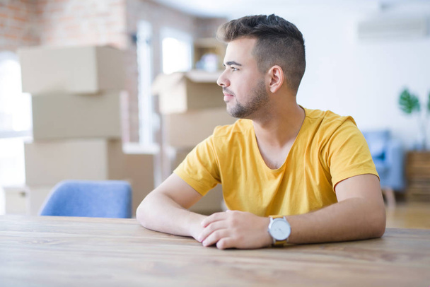 Young man sitting on the table with cardboard boxes behind him moving to new home looking to side, relax profile pose with natural face with confident smile. - Foto, Imagen