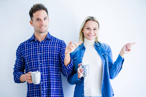 junges schönes Paar, das eine Tasse Kaffee trinkt, steht vor isoliertem weißem Hintergrund und zeigt glücklich mit der Hand und dem Finger zur Seite - Foto, Bild