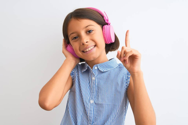 Belle enfant fille écoutant de la musique à l'aide d'écouteurs sur fond blanc isolé surpris par une idée ou une question pointant du doigt avec un visage heureux, numéro un
 - Photo, image