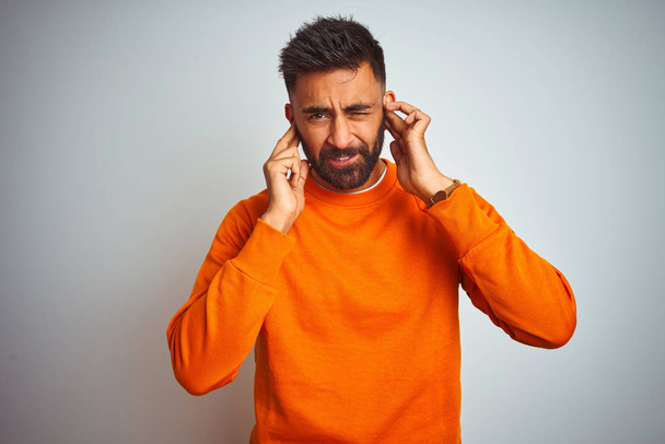 Jonge Indiase man dragen oranje trui over geïsoleerde witte achtergrond bedekken oren met vingers met geïrriteerde uitdrukking voor het lawaai van luide muziek. Doof concept. - Foto, afbeelding