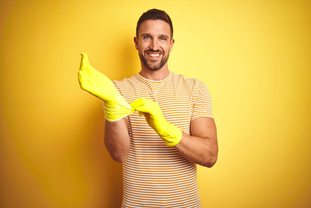 Joven hombre guapo usando guantes de limpieza para las tareas domésticas sobre un fondo amarillo aislado con una cara feliz de pie y sonriendo con una sonrisa segura que muestra los dientes
 - Foto, Imagen