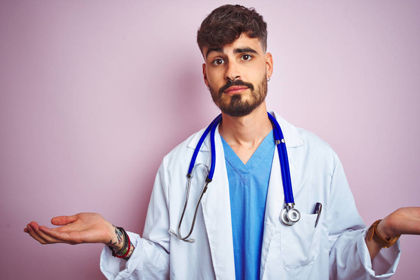 孤立したピンクの背景の上に立ってステトコペを着た入れ墨を持つ若い医者の男は、腕と手を上げて、無知で混乱した表情をしています。疑いの概念. - 写真・画像