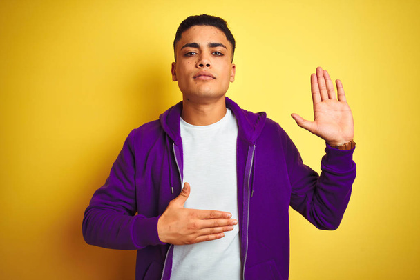 Nuori brasilialainen mies yllään violetti huppari seisoo eristetyn keltaisen taustan yläpuolella vannoen käsi rinnassa ja avoin kämmen, tehden uskollisuuslupauksen
 - Valokuva, kuva