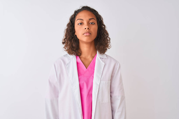 顔に真剣な表情で隔離された白い背景の上に立ってコートを着た若いブラジル人医師の女性。シンプルで自然なカメラを見て. - 写真・画像