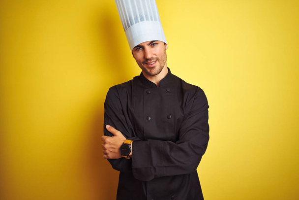 Young chef man wearing uniform and hat standing over isolated yellow background happy face smiling with crossed arms looking at the camera. Positive person. - Photo, Image