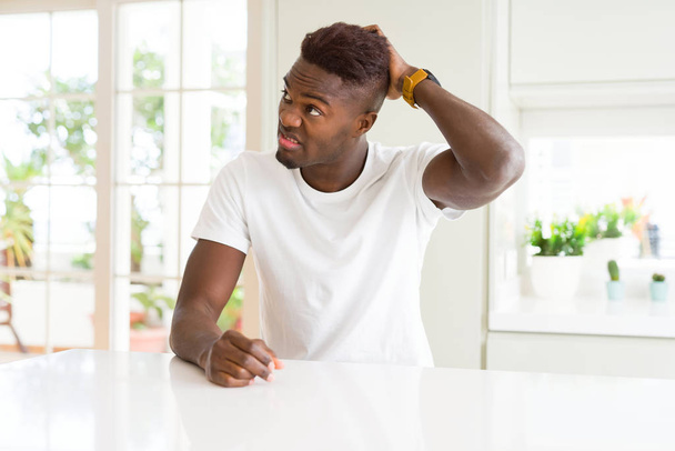 Handsome african american man on white table at home confuse and wonder about question. Uncertain with doubt, thinking with hand on head. Pensive concept. - Foto, imagen
