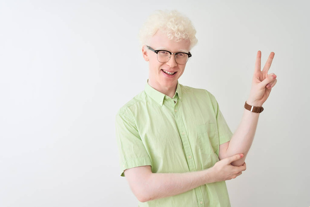Mladý albín blonďatý muž v zelené košili a brýlích na izolovaném bílém pozadí, usmívající se s veselou tváří mrkající na kameru, která dělá znamení vítězství. Číslo dvě. - Fotografie, Obrázek