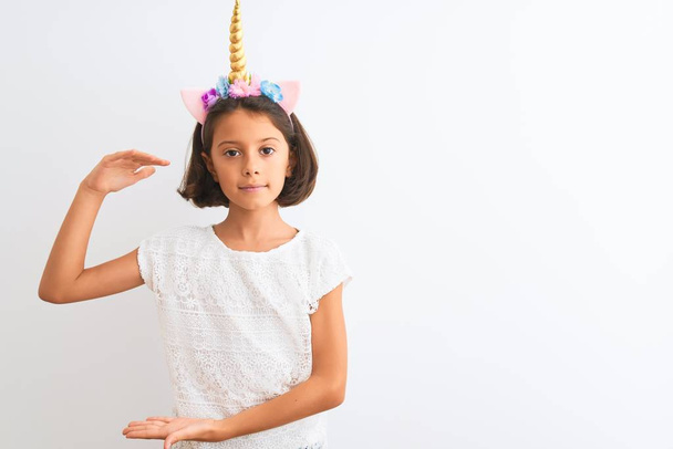Mooie kind meisje dragen Unicorn diadeem staande over geïsoleerde witte achtergrond gebaren met handen tonen groot en groot formaat teken, meet symbool. Glimlachend kijken naar de camera. Meet concept. - Foto, afbeelding