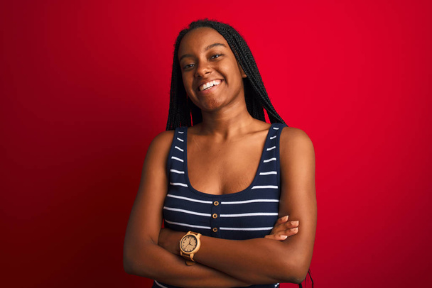 Giovane donna afroamericana che indossa maglietta a righe in piedi sopra isolato sfondo rosso volto felice sorridente con le braccia incrociate guardando la fotocamera. Persona positiva
. - Foto, immagini