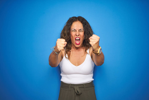 Middle age senior woman with curly hair standing over blue isolated background angry and mad raising fists frustrated and furious while shouting with anger. Rage and aggressive concept. - Photo, Image