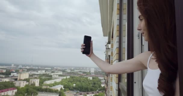 Pencereden bakan ve akıllı telefonda selfie çeken genç kadın. - Video, Çekim