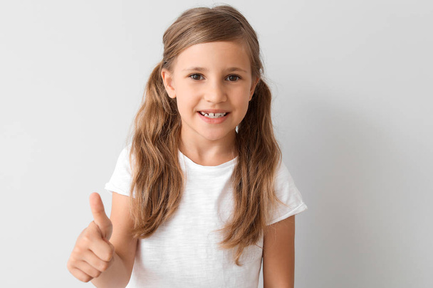 Little girl in stylish t-shirt showing thumb-up on light background - Foto, Imagem