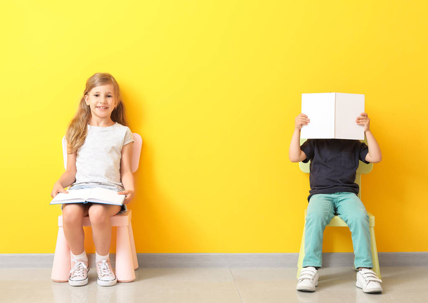 Маленькие дети с книгами сидят на стульях возле цветной стены
 - Фото, изображение