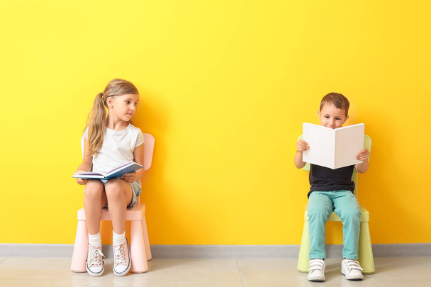 Маленькие дети с книгами сидят на стульях возле цветной стены
 - Фото, изображение