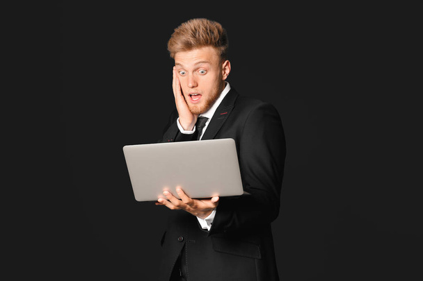 Shocked young businessman with laptop on dark background - Foto, imagen