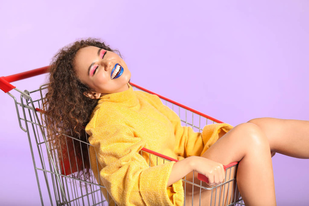 Portrait of fashionable young African-American woman in shopping cart on color background - 写真・画像