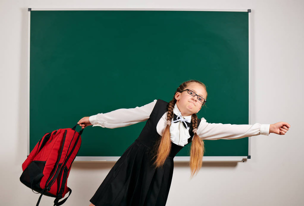 Portrait of a schoolgirl playing with backpack and school supplies near blackboard background - back to school and education concept - Photo, Image