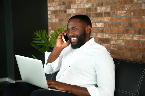 Όμορφος Αφροαμερικανός επιχειρηματίας με φορητό υπολογιστή που μιλά από το κινητό τηλέφωνο στο γραφείο - Φωτογραφία, εικόνα