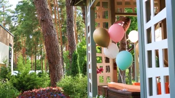 Bando de balões de ar de hélio coloridos diferentes que jogam no vento amarrado ao terraço gazebo ao ar livre de madeira como decoração para festa de aniversário de crianças. Aniversário celebração decoração design
 - Filmagem, Vídeo