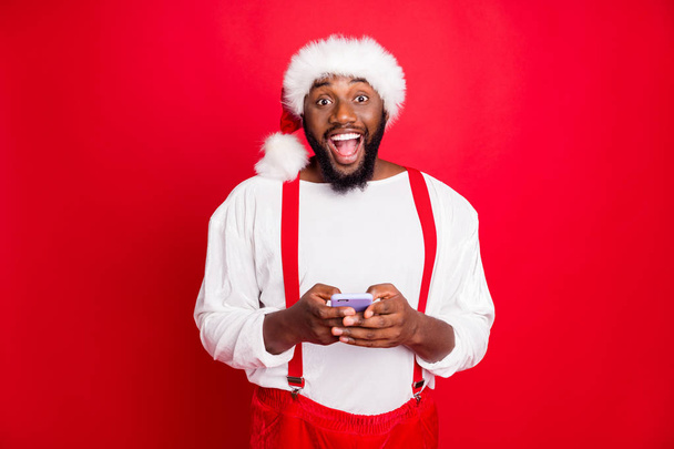Portrait of impressed black santa claus in xmas hat screaming wow omg use cell phone modern technology got noel congratulations wearing white pullover isolated over red background - Foto, immagini