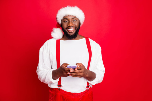 Portrait of cheerful black santa claus in xmas hat using cell phone gadget read new year posts wearing white sweater isolated over red background - Photo, image