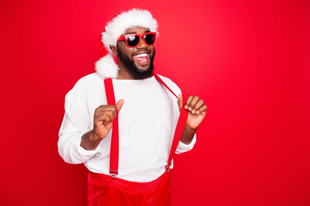 Turned photo of cheerful afro santa claus touching his suspenders posing wearing white pullover eyeglasses eyewear isolated over red background - Foto, Imagen
