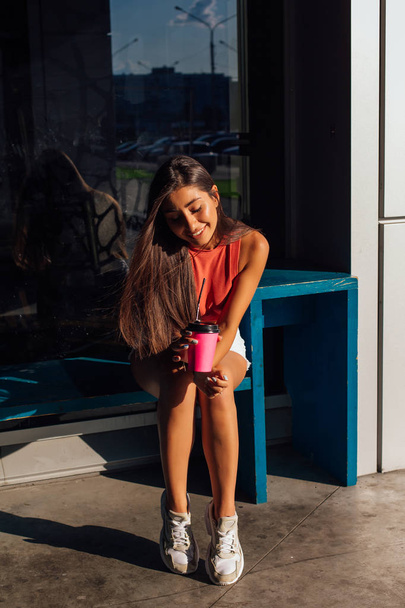 Stylish happy young brunette woman wearing white shorts and sneakers holding pink cup of coffee to go. - Фото, изображение