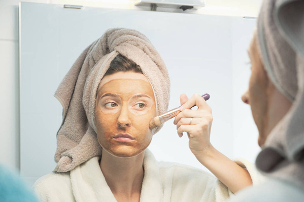 Jeune femme met masque d'argile sur son visage avec brosse regardant dans le miroir dans la salle de bain
.  - Photo, image