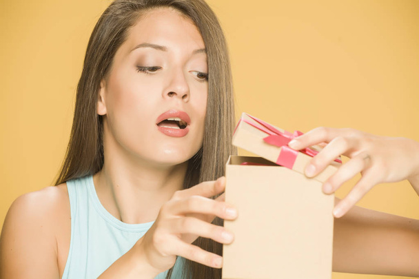 Young beautiful curious woman opening a present box on yellow background - Foto, afbeelding