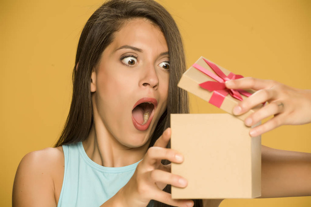 Νεαρή σοκαρισμένος γυναίκα ανοίγοντας ένα παρόν κουτί σε κίτρινο φόντο - Φωτογραφία, εικόνα